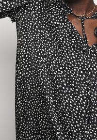 Haut en tissu noir avec un motif floral blanc, fermeture à boutons et un détail de nouage au niveau du col, avec une finition légèrement texturée.