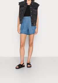 Femme portant un gilet en jean noir sans manches, un haut noir, un short en jean bleu taille haute avec ceinture, et des sandales noires, debout sur un sol gris.