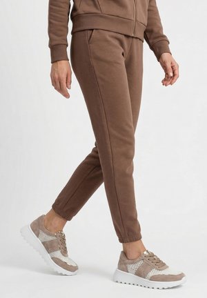 Person iført brune joggingbukser og beige hvide mesh sneakers, der står på hvid baggrund.