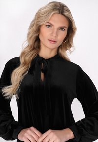 Blusa nera in velluto con scollatura a chiave, lacci al colletto e maniche lunghe a palloncino. Tessuto liscio con una finitura lucida.