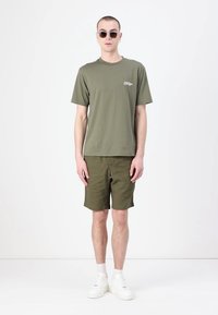 Tricou din bumbac de culoare verde măsliniu, cu mâneci scurte, logo pe piept; șorturi verzi măslinii asortate; teniși albi; ochelari de soare rotunzi; fundal simplu.