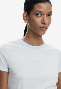 T-shirt bleu clair  à manches courtes en tissu doux, présentant un petit texte "Conçu" en cristaux brillants sur le devant.