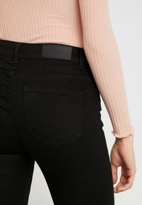 Primer plano de una persona que lleva unos pantalones vaqueros negros con un bolsillo trasero y una camiseta de manga larga de rayas en rosa claro.