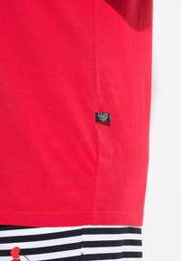 T-shirt en coton rouge avec un col rond, présentant une étiquette noire sur le côté. En dessous, un tissu à rayures noires et blanches est partiellement visible.