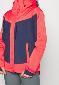 Röd och marinblå vädertålig jacka med en dragkedjeförsedd frontficka, justerbar huva och kardborrefästen. Har kontrasterande färgpaneler och textur.