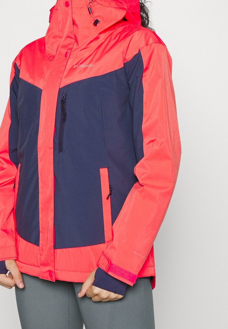 Röd och marinblå vädertålig jacka med en dragkedjeförsedd frontficka, justerbar huva och kardborrefästen. Har kontrasterande färgpaneler och textur.