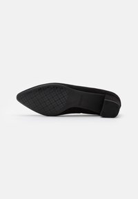 Zapato plano de ante negro con puntera afilada y suela de goma texturizada que presenta un patrón geométrico. Diseño de tacón bajo.