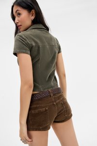 Una giovane donna con lunghi capelli neri indossa una maglietta verde oliva a maniche corte e pantaloni corti in velluto a coste marroni con cintura borchiata, rivolta di spalle con lo sguardo sopra la spalla.