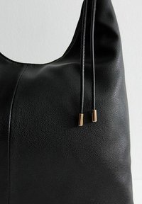 Bolso tote negro de cuero con una superficie texturizada, que presenta dos cordones ajustables que terminan en puntas metálicas de color dorado.