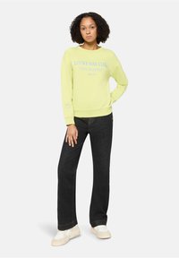 Jasnożółta bluza z napisem "LUCKY DAY CLUB", miękki materiał, długie rękawy, ściągacze na mankietach, w połączeniu z czarnymi dzwonami i jasnymi sneakersami.