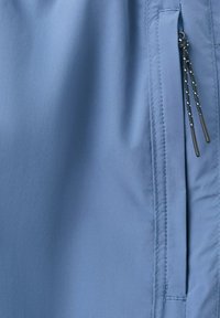 Gros plan d'un tissu bleu clair avec une couture verticale et une poche avec deux cordons gris et blancs aux extrémités en caoutchouc.