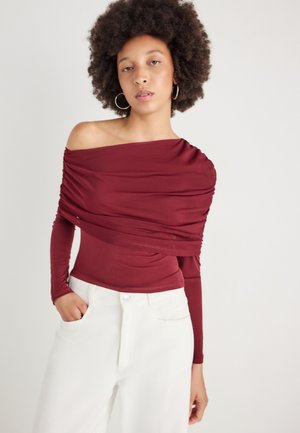 Top bordeaux à épaules dénudées avec des manches longues, présentant des détails de tissu froncé et une texture lisse, associé à un pantalon blanc taille haute.
