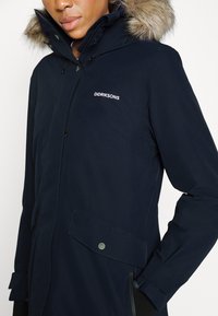 Námořnická modrá parka s kapucí s kožíškem, předním zipem a patentkami, postranními kapsami a logem "DIDRIKSONS" na hrudi.