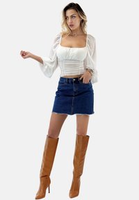 Weißes langärmliges Oberteil mit gerafft Vorderseite, kombiniert mit einem dunkelblauen Denim-Minikleid. Getragen mit tanfarbenen kniehohen High Heels.