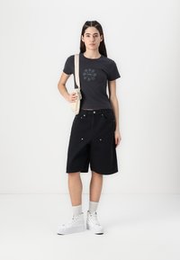 Maglietta grigia con un design ricamato circolare, abbinata a shorts di jeans neri. Sneakers bianche e una borsa a tracolla color crema completano il look.