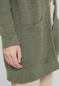Olivengrøn strikket cardigan med struktureret overflade, ribbede manchetter og en lomme foran. Stoffet er blødt med en afslappet pasform og sideskårne slids.
