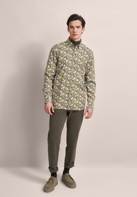 Blumenmuster-Hemd in Weiß, Gelb, Blau und Grün mit langen Ärmeln; kombiniert mit olivgrünen Hosen und beigen Schuhen.