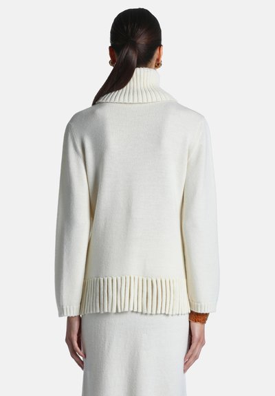 Femme aux cheveux foncés en queue de cheval portant un pull à col roulé côtelé blanc et une jupe assortie, debout de dos avec un poignet marron visible.