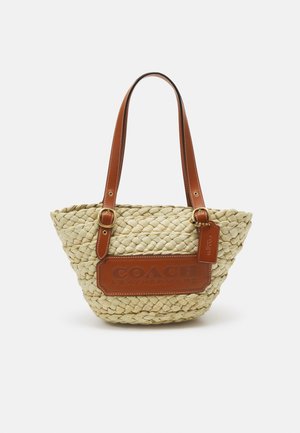 Sac cabas en paille tressée avec poignées en cuir beige et patch logo, avec une petite étiquette en cuir, sur fond blanc uni.