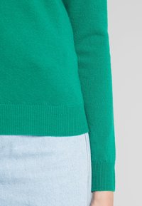 Maglione verde lavorato a maglia con polsini e orlo a costine. La texture appare morbida e viene abbinato a un pantalone in denim chiaro.