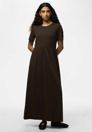 Vrouw met lang krullend haar, gekleed in een donkerbruine maxi-jurk met korte mouwen en zwarte schoenen, staand met haar handen achter haar rug.