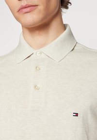 Polo beige con colletto classico, tre bottoni e un piccolo logo ricamato in blu navy, rosso e bianco sul lato sinistro del petto. Tessuto textured.