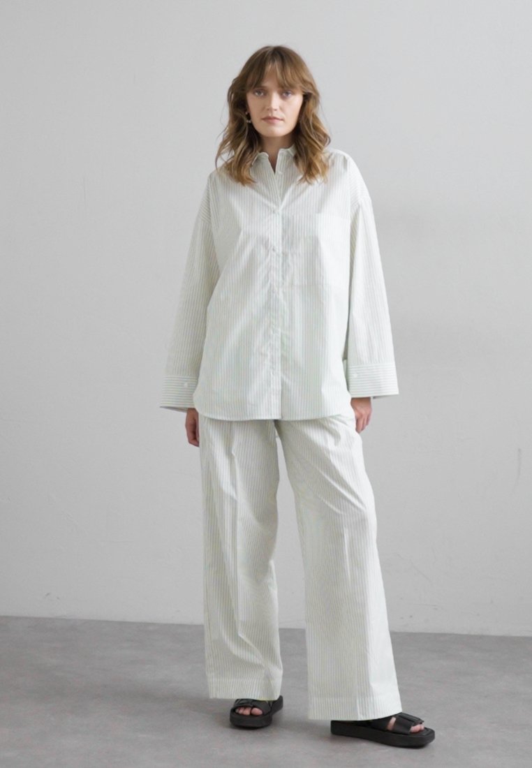 Chemise surdimensionnée rayée vert clair et blanc à manches longues, associée à un pantalon large assorti et des sandales noires. Coupe simple et décontractée.