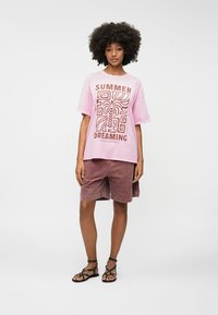 Roza oversize majica s smešnim smeškom u smešnom tonu koja se čita "SUMMER DREAMING", uparen s burgundskim šortsem i crnim sandalama na remenčiće.