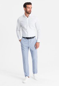 Camicia bianca con piccoli pois blu navy, abbinata a pantaloni azzurri chiari, cintata con una cintura scura. Le scarpe sono sneakers bianche.