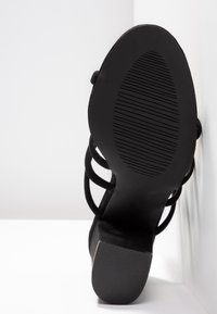 Sandale noire à talon avec plusieurs lanières, vue de la semelle, posée contre des surfaces d’angle blanches.