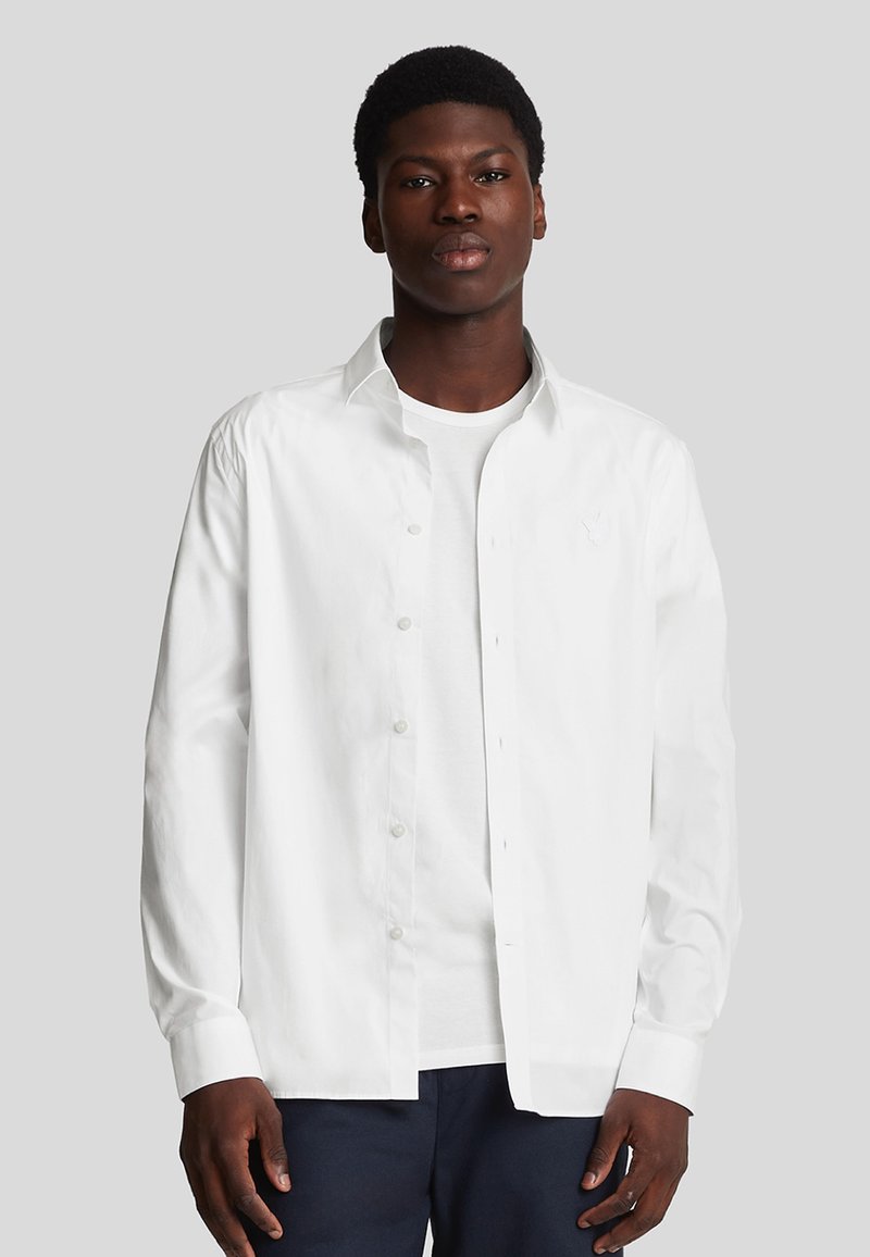 Camicia bianca a maniche lunghe con bottoni frontali, tessuto morbido, vestibilità rilassata e logo ricamato sul petto. Indossata sopra una maglietta bianca.