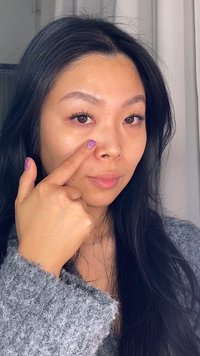 Modell mit langen schwarzen Haaren, das einen strukturierten grauen Pullover trägt und mit lila glitzerndem Nagellack auf ihre Nase zeigt. Natürliches Make-up, neutraler Hintergrund.