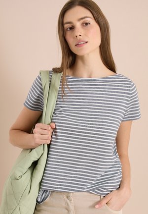Ung kvinde iført en grå og hvidstribet T-shirt, der holder en lys grøn quiltet jakke over den ene skulder mod en beige baggrund.