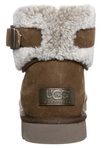 Bruine suède enkellaars met grijze schapenvachtvoering, metalen gespsluiting en een geperst UGG-logo op de hiel. Platte rubberen zool.