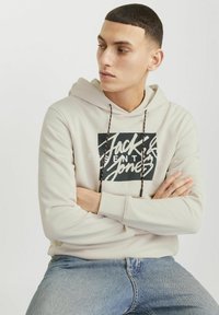 Lys beige hoodie med sort grafikprint, justerbar snor med rød og sort accent, parret med jeans i blå denim.
