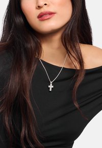 Silberne Kreuz-Halskette an einer zarten Kette, getragen �über einem schwarzen schulterfreien Oberteil, mit glatten Texturen und einem minimalistischen Design.