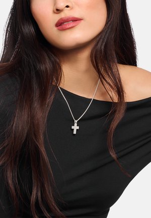 Silberne Kreuz-Halskette an einer zarten Kette, getragen über einem schwarzen schulterfreien Oberteil, mit glatten Texturen und einem minimalistischen Design.