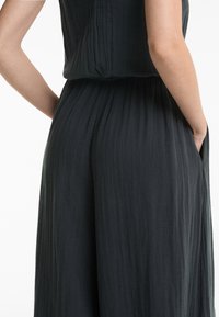 Temno siva brezrokavna kombinezona iz teksturirane, gubane tkanine, ki se ponaša z udobnim krojem in stranskimi žepi, ter nabrano podrobnostjo na pasu.