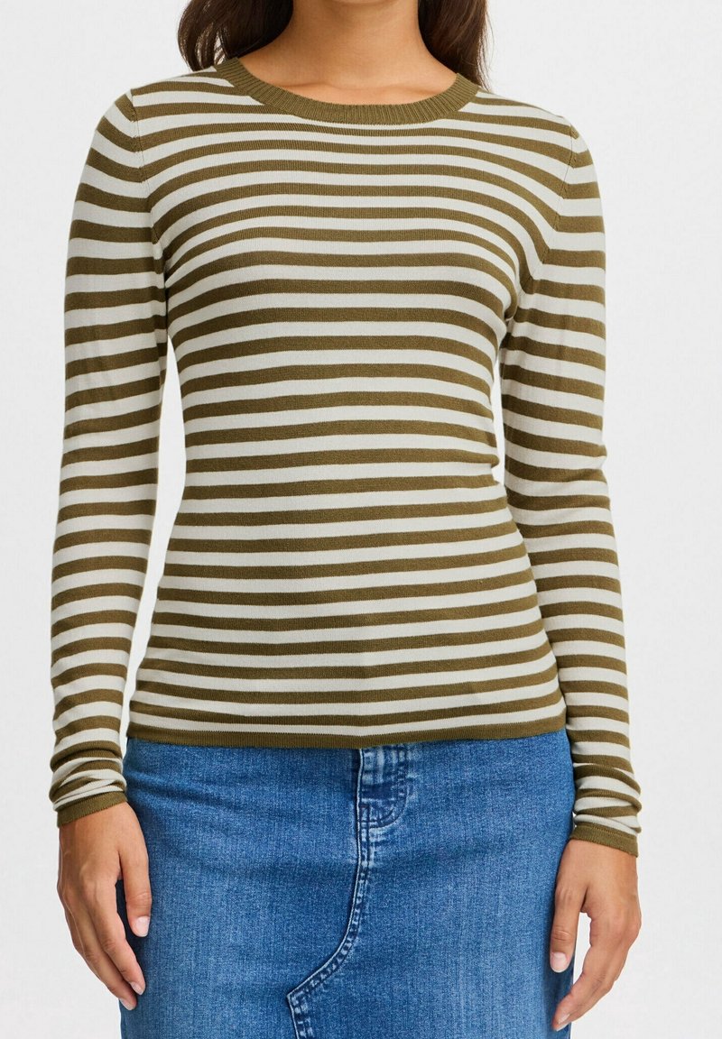 Femme portant un t-shirt à manches longues à rayures horizontales vert olive et blanc, et une jupe en denim bleu, debout avec les mains détendues.