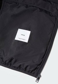 Intérieur de veste noire avec une fermeture éclair et une étiquette d'entretien portant le logo "MNG". Texture lisse avec un léger éclat. Les coutures sont visibles.