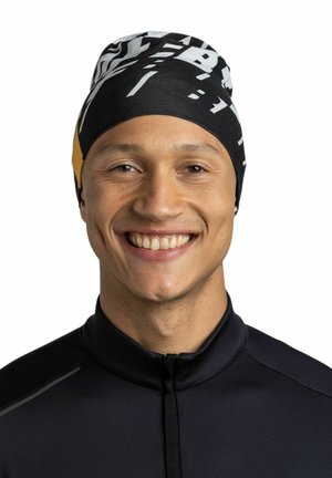 Black and yellow beanie with bold white graphic patterns, fitted design, smooth texture. Worn by a smiling individual in a dark zip-up top.