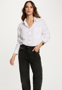 Camisa blanca de botones con dos bolsillos en el pecho, con un acento de cuello texturizado. Combinada con unos vaqueros negros de cintura alta.