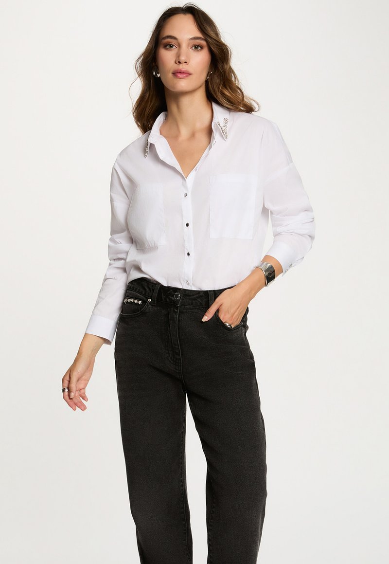 Camisa blanca de botones con dos bolsillos en el pecho, con un acento de cuello texturizado. Combinada con unos vaqueros negros de cintura alta.