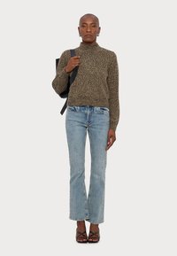 Pull en maille texturée marron avec un col montant, associé à un jean droit bleu clair et des sandales noires à lanières. Sac à dos noir porté sur l'épaule.