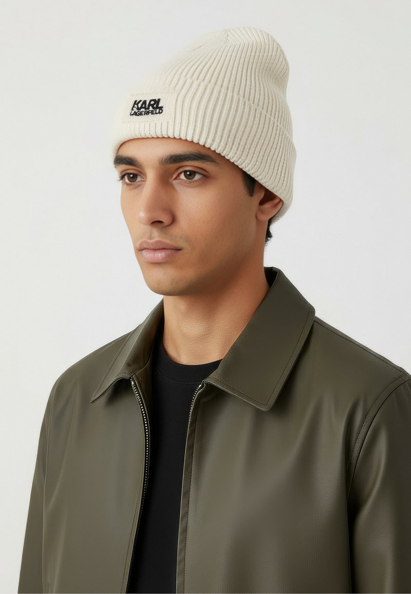 Off-white ribbed knit beanie with black logo patch. Model wears an olive green jacket over a black shirt, featuring a classic collar and zipper.