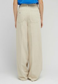 Beżowe spodnie typu wide-leg wykonane z bawełny, z dwiema tylkimi kieszeniami o zaokrąglonych szwach oraz małą metką z logo na prawej kieszeni.