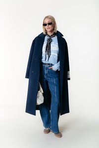 Abrigo navy sobre un sudadera azul claro con texto blanco, jeans de pierna ancha con patrón de rayas, gafas de sol y una bolsa de malla.