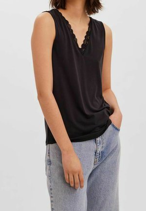 Vrouw draagt een mouwloze zwarte top met kanten rand en lichtblauwe jeans met hoge taille, staat met één hand in de zak.