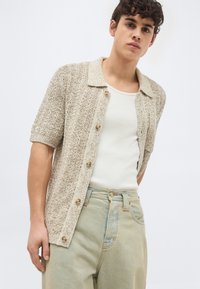 Giovane uomo che indossa una camicia beige a maniche corte con trama sopra una canottiera bianca e jeans a vita alta dall'aspetto slavato, in piedi davanti a uno sfondo semplice.