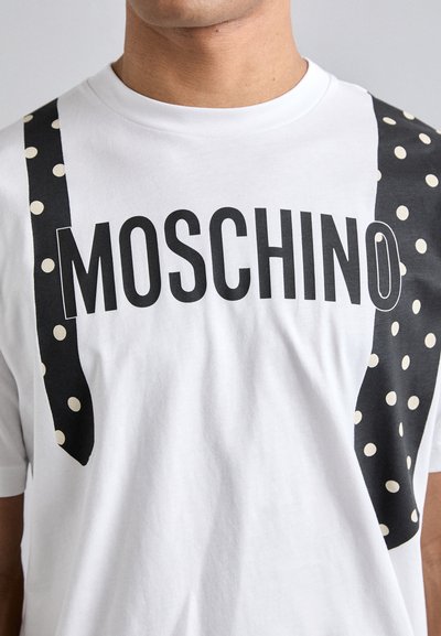 Balta kokvilnas T-kreklu ar izteiksmīgu melnu "MOSCHINO" logotipu. Melni akcenti ar baltām punktiņām uz pleciem, radot piekareņu efektu.